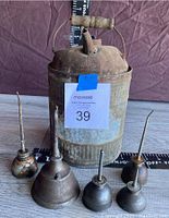 Photo showing 4 quart metal oil can with wooden handle and five vintage thumb oilers arranged in front on a wood surface with measuring tape for scale.
