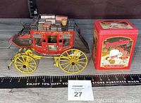 Side front angle of Wells Fargo stagecoach next to Burma-Shave gift tin showing detailed model with luggage on roof and tin's colorful advertising graphics.