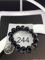 View of large black agate beaded stretch bracelet laid on a reflective surface alongside a Canadian quarter for size comparison.