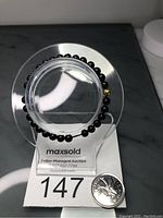 Black Burma jade stretch bracelet displayed on clear circular stand with auction lot label and coin for scale.