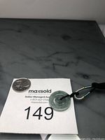 Close up photo of Burma jade double round pendant on adjustable black rope with metallic and turquoise beads, placed on a numbered auction card and surface.