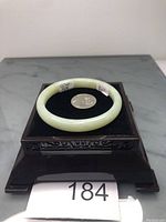 Light green jade bangle bracelet on black velvet cushion with coin for scale, displayed on carved dark wooden stand.