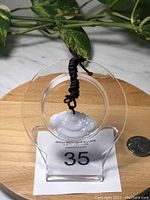 Front view of the white Burma jade wish-bone-shaped pendant displayed on a clear stand with black adjustable braided rope necklace, placed on a wooden surface beside a quarter for scale.