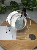 Light green jade pendant displayed on a clear circular stand, with black adjustable rope and red/green beads, quarter placed nearby for size.
