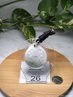 Full view of round jade pendant displayed on a clear acrylic stand with a black woven adjustable rope and a quarter coin for size comparison on a wooden surface.