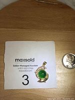 Green circular gemstone pendant with gold-tone decorative setting and bail. Shown with coin for scale.