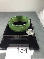 Polished green agate stone bangle bracelet displayed on a decorative stand next to a quarter for scale, showing the rounded band shape, varied green colors and natural stone patterns.