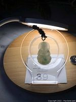 Front view of translucent green Burma jade Kwan Yin Buddha pendant on adjustable black braided rope, displayed on acrylic stand with auction lot number visible.