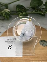 Front view of Burma jade Money God pendant with visible natural grey jade inclusions and silver clip, placed on a clear display stand next to a coin for size reference.