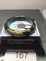 Dark green nephrite jade bangle with gold calligraphy painted decoration, displayed on wooden stand with testing license and coin for scale.