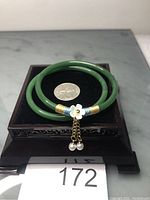 Photo of two green jade bangles on a black display stand with a quarter for scale. One bangle features a small white flower charm with gold chains and white pearl-like dangles wrapped with gold and blue thread. The other bangle is plain green jade.