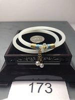 Pair of white jade bangles with flower and pearl dangle, wrapped in blue and gold thread, shown on a black stand with a coin for size reference.