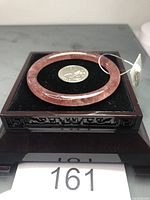 Photo of the strawberry quartz bangle displayed on a black cushion inside a carved wooden box, with a coin for scale and lot tag.