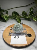 Front view of the round light green Burma jade pendant featuring a tiger design, displayed on a clear stand with a token coin for size reference.