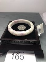 Tourmaline bangle bracelet displayed on a decorative black stand against a neutral background. The bangle shows natural color variations with white, light gray, purple, and pink hues. A quarter coin is placed inside the bangle for scale. A small white price tag is attached.