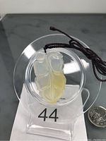 Front view of the carved yellow and white gemstone pendant on an adjustable dark rope, placed on a clear stand with a quarter coin for size reference.