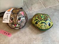 Photo of two round painted rocks placed on the floor, one painted as a cat face and the other as a frog face, showing detailed painting and color patterns.