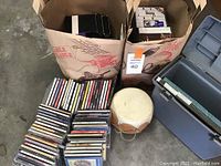 Photo showing paper bags filled with CDs, a wooden bongo drum, and a gray plastic file box with a handle