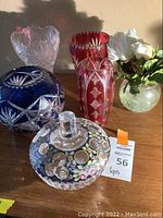 Photo showing five decorative colored glass items including a cobalt blue etched vase, two tall cut glass vases in red and clear, a floral covered jar, and a small clear vase with faux white flowers.