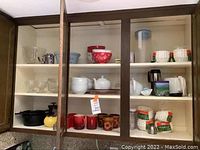 Cabinet interior showing a collection of assorted bowls, teapots, glassware, mugs, and coffee filters on shelves.