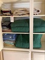Shelves filled with folded clean linens and towels, including green quilted blankets, blue bath towels, and assorted towels in cream, gray, maroon, tan, and green colors.