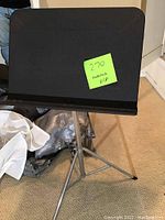 Metal music stand with black desk surface and collapsible silver tripod base setup on carpeted floor.