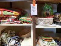 Shelf with colorful folded outdoor plastic coated tablecloths, woven bread baskets, and stacked napkins.