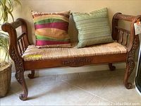 Full front view of the wooden bench with carved detailing and two decorative pillows on the wicker seat.