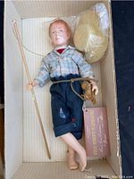 Photo of the Huckleberry Finn doll lying in a white striped box holding a fishing pole, wearing a plaid shirt and patched pants, with a straw hat included.