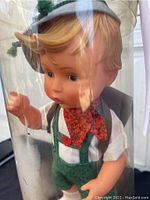 Close-up of the Goebel doll's face and upper body inside the clear plastic cylinder, showing detailed facial features, blond hair, and traditional clothing with suspenders and neck scarf.