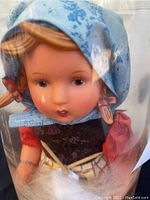 Close-up front view of the Goebel doll with blue headscarf, peeking through original plastic cylinder packaging.