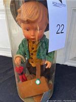 Front view of the Goebel doll inside the original clear plastic cylinder. Doll has blonde hair, green jacket, brown vest, and black pants. Accessories visible include a brown satchel and a red item.