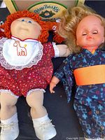 Photo showing two dolls lying next to the original Cabbage Patch Kids box. One doll is a Cabbage Patch Kids doll, the other a Japanese doll in kimono.
