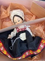 Full view of 7-inch Madame Alexander doll wearing traditional dress with embroidered skirt, lace bonnet, seated in original box with orange tissue paper lining