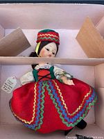 Image showing the Madame Alexander doll in traditional Czech dress lying in the original pink cardboard box with divider, doll wearing red skirt with multicolor rickrack, head detached inside the box.