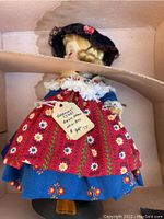 Side view of the doll inside the original box showing the dress, hat, and label attached to the doll.
