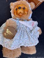 Raikes Susie Bear in blue floral dress with lace trim, bonnet, wooden face, plush fur body, and tag on arm.