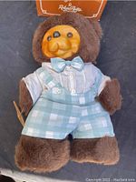 Full body of the Raikes Timmy Bear plush with wooden face dressed in light blue shirt, bow tie, and checkered overalls.
