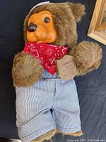 Raikes Bear stuffed toy wearing red bandana and striped overalls, showing brown fur, orange plastic face, and a tag attached to its hand.