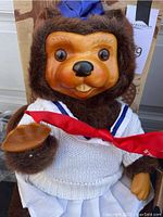 Close-up of Raikes Originals brown bear figure wearing white sweater and red scarf, showing detailed sculpted face and plush fur limbs.