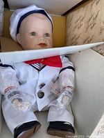 Doll seated inside original box, dressed in white sailor suit with red bow and plastic-covered hands.
