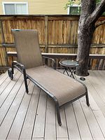 Metal framed chaise lounge with brown woven fabric seating and wheels on back legs, placed on a deck near wooden fence.