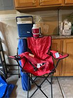 Red folding camp chair with carry bag and cooler with two thermoses on top, flashlights also present