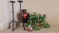 Two tall iron candle holders, faux green plant in woven basket, metallic red decorative vase, and small copper-colored candle tin displayed on shelf