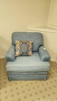 Front view of blue denim armchair with decorative pillow on beige patterned carpet.