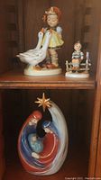 Photo showing three porcelain figurines: two Goebel Hummel figures (girl with geese and boy on fence) and one nativity scene.