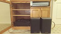 Wooden cabinet opened showing Kenwood stereo receiver and Magnavox DVD/VHS player on top shelf, Polk Audio speakers beside cabinet.