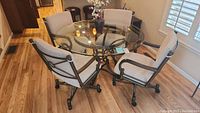 View of round glass top table with four gray cushioned rolling chairs arranged around it on wood floor.