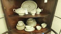 Full set displayed on two shelves inside a corner cabinet showing the soup tureen, platter, plates, cups, saucers, creamer and sugar bowl, highlighting overall quantity and appearance.