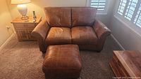 View of loveseat and ottoman set in a carpeted room next to an end table and lamp.